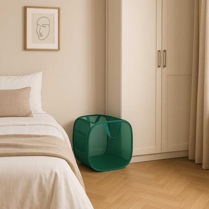 Sammenklappelig Vasketøjskurv i Mesh - Let, Pladsbesparende i Mørkegrøn og Sort. A stylish bedroom setting with the foldable green laundry basket neatly tucked beside a wardrobe, highlighting its space-saving feature and ease of storage in a minimalist decor environment.