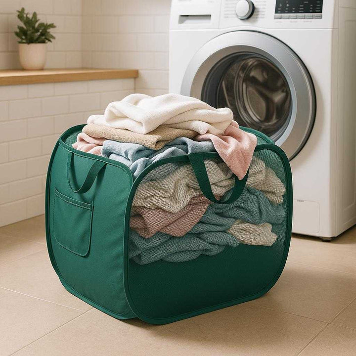 Sammenklappelig Vasketøjskurv i Mesh - Let, Pladsbesparende i Mørkegrøn og Sort. The collapsible mesh laundry basket sits filled with freshly washed clothes in a brightly lit laundry room, showcasing its spacious yet compact design and dark green color against a modern washing machine backdrop.