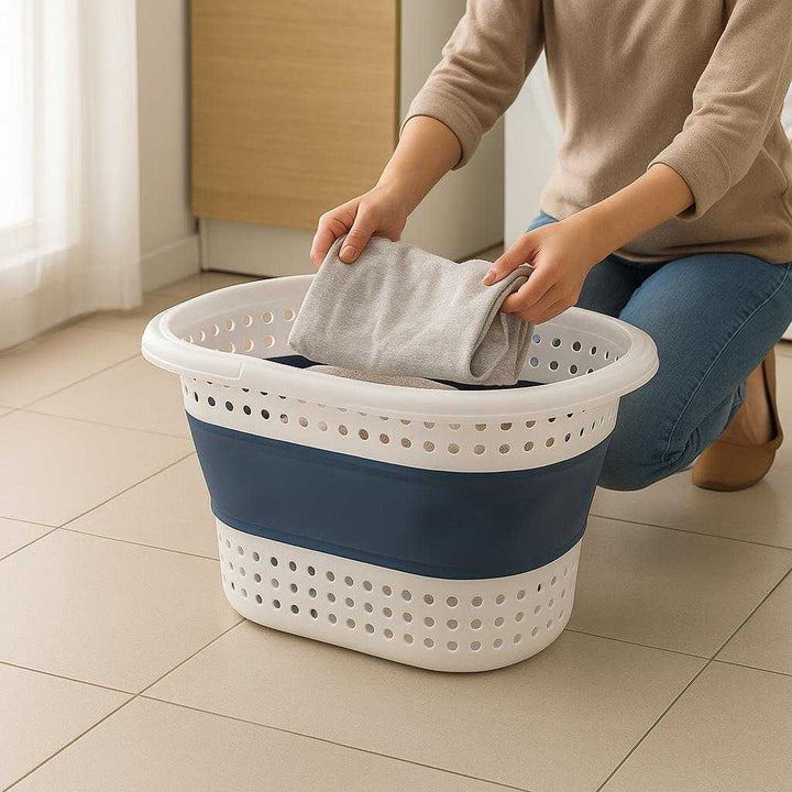 Sammenklappelig vasketøjskurv i plast og silikone – Let, rummelig og pladsbesparende i blå og hvid: A realistic laundry room setting where the collapsible laundry basket is properly grounded on a clean tiled floor, being actively used by a person sorting freshly laundered clothes into it.