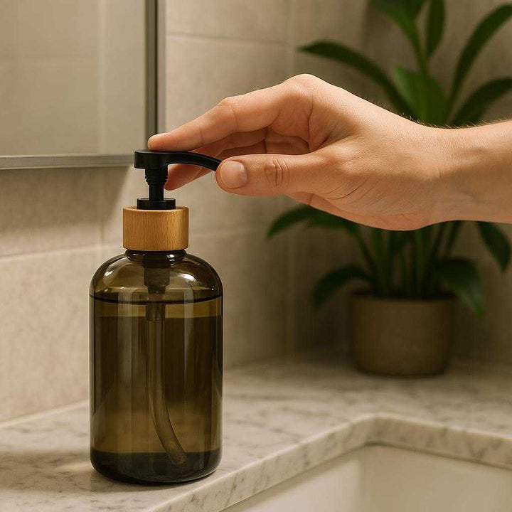 Sæbedispenser i Røgfarvet Glas med Sort Pumpe - 500ml Høj Kvalitet og Stilfuldt Design: A realistic bathroom setting where the soap dispenser is properly grounded on a marble sink counter, actively used by a person’s hand pressing the pump, with natural lighting casting realistic shadows and showing clear contact points where the dispenser and counter meet.