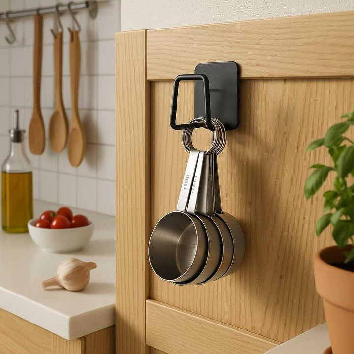 Selvklæbende vægkrog i mat sort metal - Moderne og pladsbesparende ophæng til hjemmet: A different realistic kitchen setting where the selvklæbende vægkrog i mat sort metal is securely positioned on a wooden cabinet door, neatly holding a set of stainless steel measuring cups.