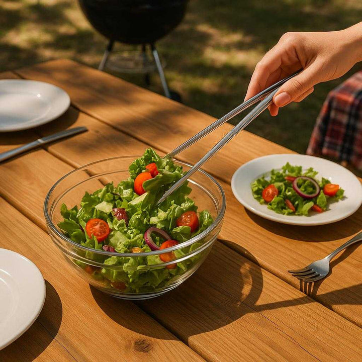 Serveringstang i Rustfrit Stål - Lille, Praktisk og Elegant i Guld Finish: A different realistic outdoor barbecue setting where the tongs are securely positioned on a picnic table, being used differently to serve salad from a large bowl, surrounded by plates and cutlery in a believable way, with proper physics, shadows from trees creating a warm ambience, and realistic integration into the casual gathering environment.