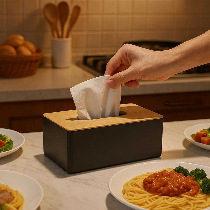 Servietboks i moderne design - Praktisk og æstetisk opbevaring til servietter i sort, lyserød, mørkeblå og hvid: A different realistic kitchen setting where the serviet box is securely positioned on a marble countertop, being actively used by a hand pulling out a napkin during a family meal, with warm ambient lighting creating distinct shadows and perfect integration into the organized kitchen environment.