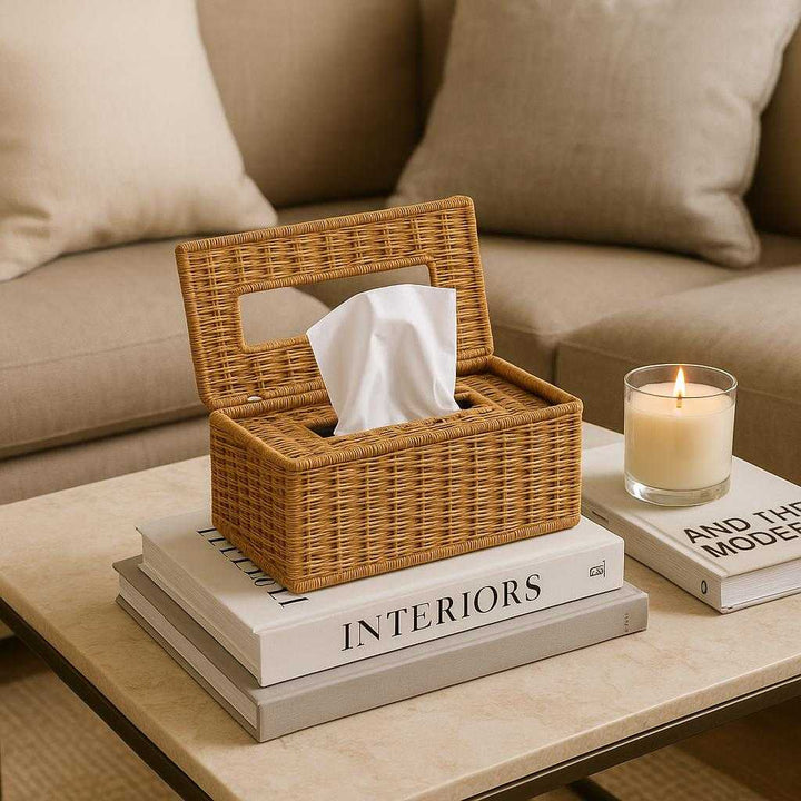 Servietæske i Flettet Rotting: Praktisk og Elegant Opbevaring til Hjemmet. The woven rattan tissue box is placed on a chic living room coffee table, surrounded by books and a lit scented candle, with a cozy sofa in the background.