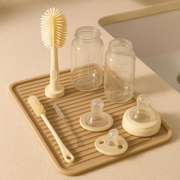 Silikone Flaske Børste - Beige & Ny Grøn, Plastik & Silicone, Udskiftelige Børstehoveder: A different realistic bathroom sink setting where the Silikone Flaske Børste is naturally placed next to a group of baby items like pacifiers and bottles on a drying mat.