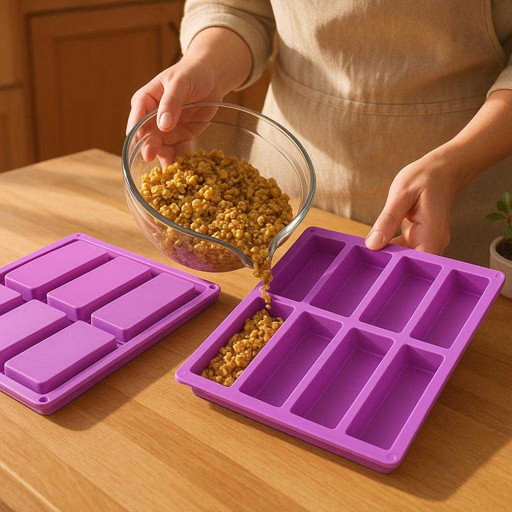 Silikone 'granola bar" → "müslibar' = 'Granola-bar' forme, lilla, fleksibel, non-stick, tåler høje temperaturer, 2 stk.: A realistic kitchen setting where the product is properly grounded on a wooden kitchen countertop, being actively used by a person to pour homemade granola mixture into the silicone mold.