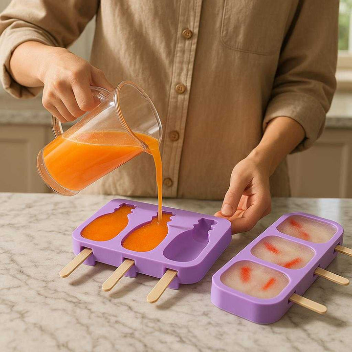 Silikone ispind form - Lilla A, B, C, D - Genanvendelig, Fleksibel: A realistic kitchen countertop setting where the product is properly grounded on the marble counter, being actively used by a person pouring fresh fruit juice into the molds, with natural lighting from a nearby window casting realistic shadows and showing clear contact points where the product touches the surface.