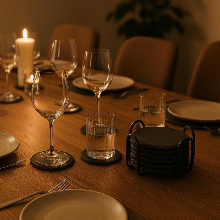 Skiferbordskånere i Sort Stålholder - Høj Kvalitet Sæt af 6 med Rustik Finish. A cozy dining room table set for a dinner party displays the Skiferbordskånere coasters in use, protecting the wooden surface from a beautifully arranged set of wine glasses and water tumblers, with ambient lighting casting gentle shadows.