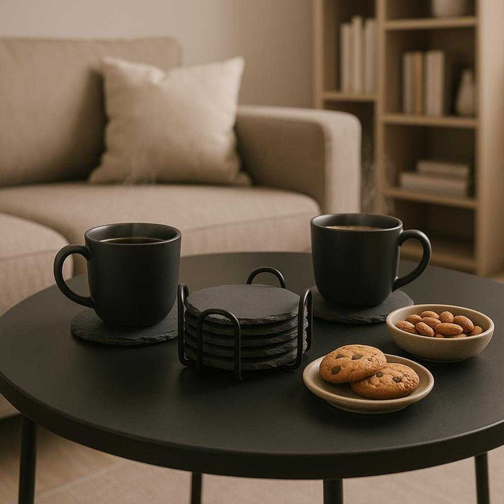 Skiferbordskånere i Sort Stålholder - Høj Kvalitet Sæt af 6 med Rustik Finish. The Skiferbordskånere coasters are styled elegantly on a modern coffee table, holding steaming cups of coffee and assortments of snacks, with a soft sofa and a bookshelf in the background showcasing a relaxing living room atmosphere.