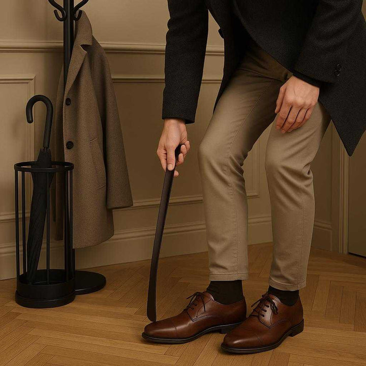 Skohorn i Sort Stål - Langt, Elegant og Funktionelt Design til Hverdagsbrug. In a stylish entry hallway, a person uses the Skohorn i Sort Stål to elegantly slide on a pair of leather shoes, with a coat rack and umbrella stand nearby.