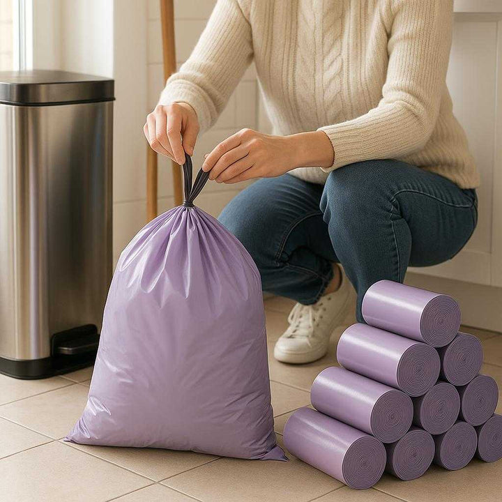 Skralde poser - Lilla, Plastik, Stor Kapacitet, Holdbarhed, Let at binde, 1 Rulle Med 100 stk, Standard Kvalitet: A realistic kitchen setting where the product is properly grounded on a tiled floor, being actively used by a person who is tying a full purple garbage bag near a stainless steel trash can with other kitchen tools like a broom and dustpan in the background.