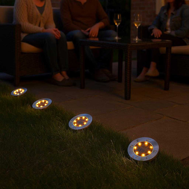 Solcellelamper til Haven: 4-pak Jordspyd med LED & Automatisk Opladning - Tilgængelig i Hvid og Varm Hvid: A different realistic backyard setting at dusk where the solar lights are placed along the edge of a patio, providing ambient light in a seating area where a small gathering is taking place.