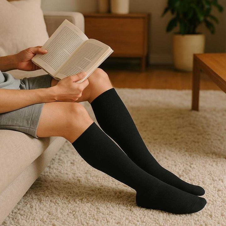 Støttestrømper i Kompression - Forbedret Blodcirkulation og Komfort i Høj Kvalitet, Sorte, Knæhøje, XS/S til XLL/XXL: A realistic home living room setting where the compression socks are properly grounded on a cozy carpet, being worn by a person relaxing on a sofa while reading a book.