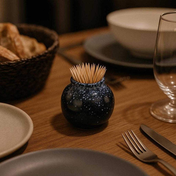 Tandstikkekrukke i keramik - Moderne og funktionel med glansfuld finish, Tilgængelig i Sort, Hvid, Brun. The ceramic holder is placed on a sophisticated dining table, filled with toothpicks, alongside minimalistic tableware and a rustic breadbasket, setting the stage for an intimate dinner gathering.