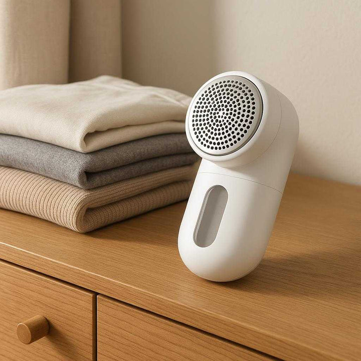 Tøjshaver med Effektiv Fnullerfjernelse - Genopliv Dine Tekstiler med Et Rent Snit i Hvid og Sølv. Displayed on a chic wooden dresser, the Tøjshaver med Effektiv Fnullerfjernelse is artfully arranged beside a stack of freshly folded clothes, sunlight highlighting its sleek white and silver design.