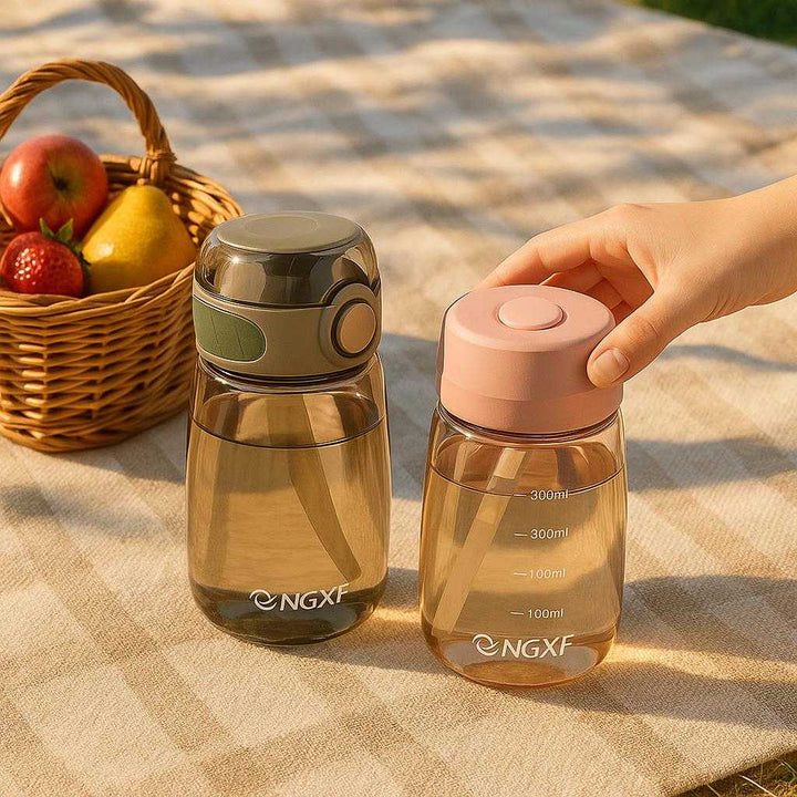 Vandflaske i Plastik og Silikone - Genanvendelig og Let at Bære - 400ml Pink, Grøn, Kaffe: A realistic outdoor picnic setting where the product is properly grounded on a picnic blanket, being naturally placed next to a small basket of fresh fruits with a person's hand casually reaching to pick it up, with natural sunlight casting realistic shadows and showing clear contact points where the product touches the blanket.