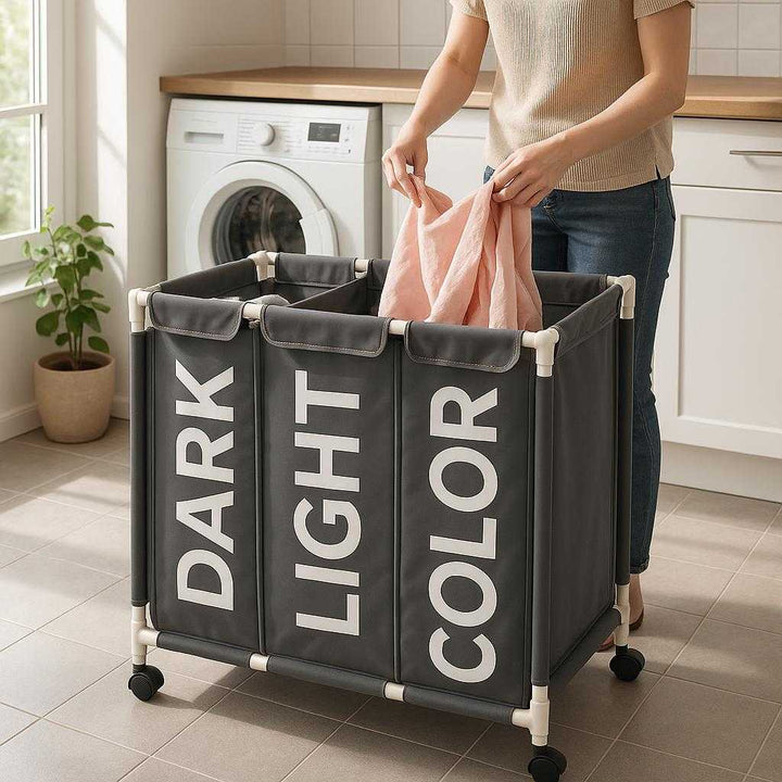 Vasketøjskurv med Tre Rum til Optimal Sortering - Professionel Grå Design med Hjul og Holdbart Stof: A realistic modern laundry room setting where the 'Vasketøjskurv med Tre Rum' is properly grounded on a tiled floor, being actively used by a person sorting laundry into its three labeled compartments.