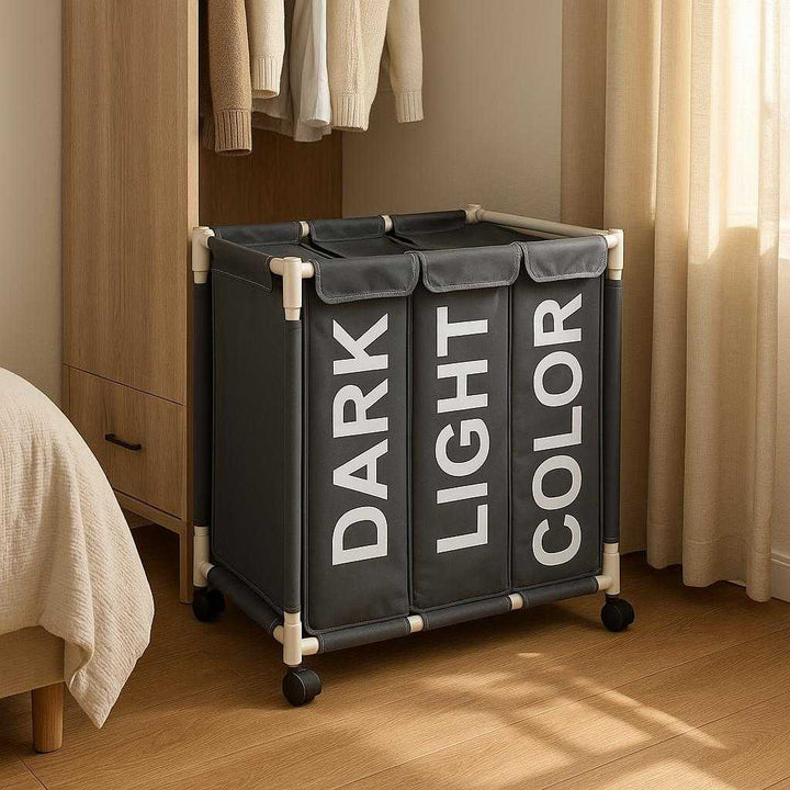 Vasketøjskurv med Tre Rum til Optimal Sortering - Professionel Grå Design med Hjul og Holdbart Stof: A different realistic bedroom corner setting where the laundry basket is securely positioned next to a wardrobe on a wooden floor, being naturally placed as part of an organized clothing and storage area.