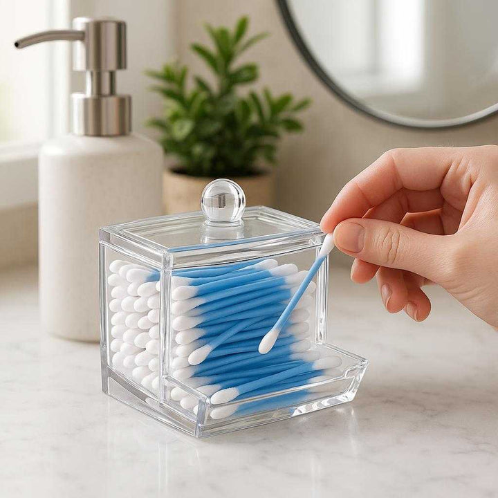 Vatpindeholder i gennemsigtig akryl og bambus - Moderne og praktisk opbevaring til badeværelset: A realistic bathroom vanity setting where the transparent acrylic and bamboo cotton swab holder is properly grounded on a marble counter, being accessed by a hand reaching for a cotton swab.