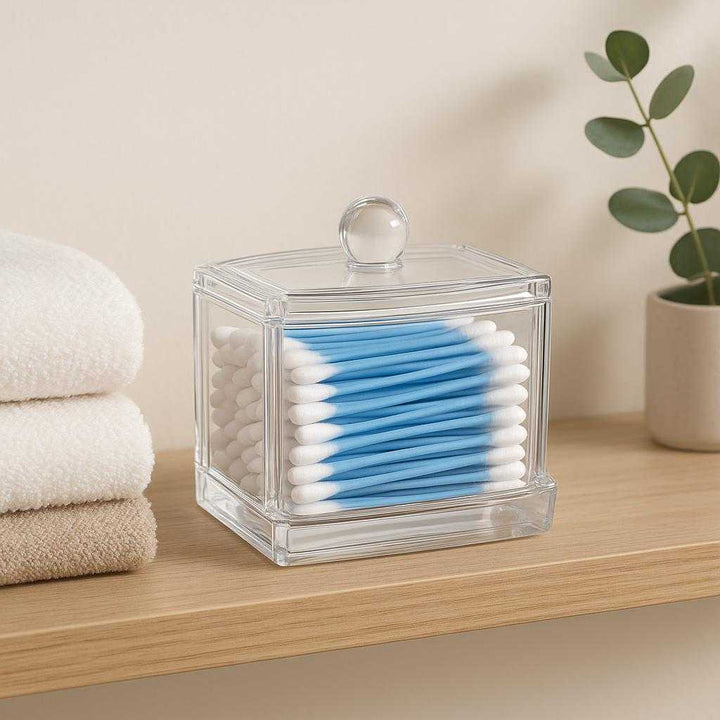 Vatpindeholder i gennemsigtig akryl og bambus - Moderne og praktisk opbevaring til badeværelset: A different realistic setting in a modern minimalist bathroom where the cotton swab holder is securely positioned on a wooden shelf, naturally placed alongside folded towels and a small eucalyptus plant.