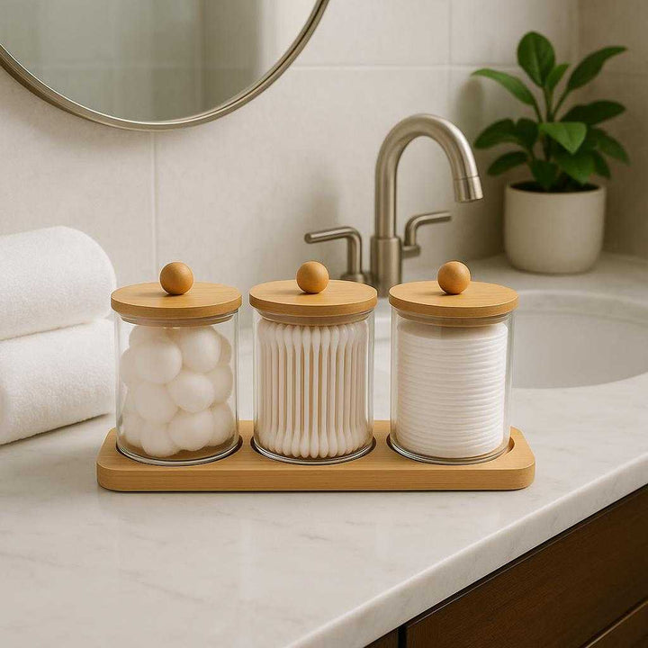 Vatpindeholdere i Glas med Bambuslåg og Bakke – Høj Kvalitet 3-delt Sæt. The glass cotton holder set with bamboo lids and tray is elegantly displayed on a bathroom counter beside a chic sink and mirror, with rolled towels and a potted plant enhancing the serene spa-like atmosphere.
