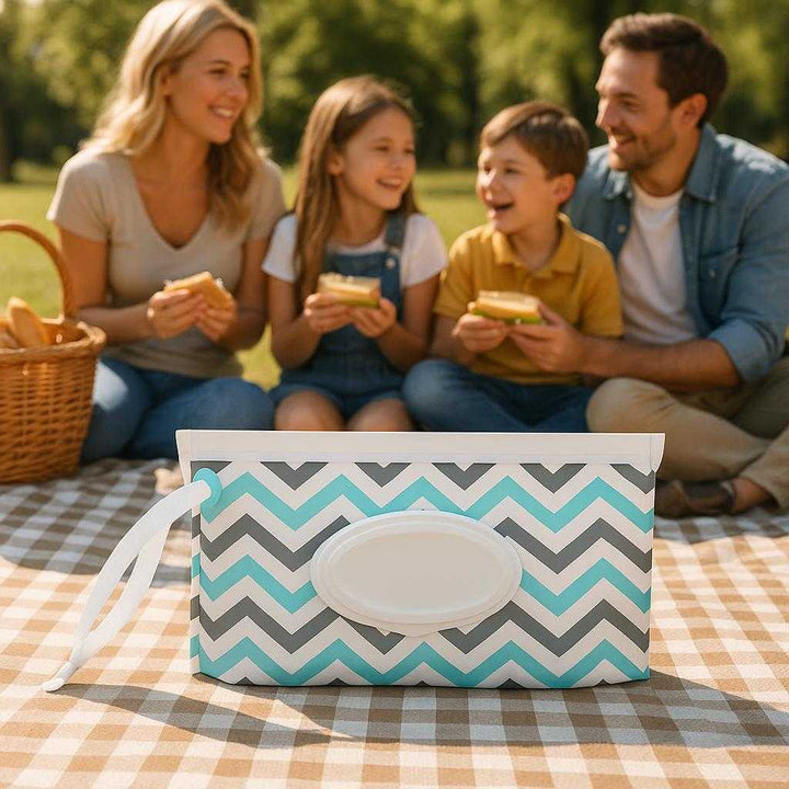Vådservietetui i Hvid og Sort - Praktisk Opbevaring med Lynlås og Dispenser. The chevron patterned wet wipe case is placed on a picnic blanket in a sunny park, surrounded by a family enjoying a picnic with a basket and sandwiches.