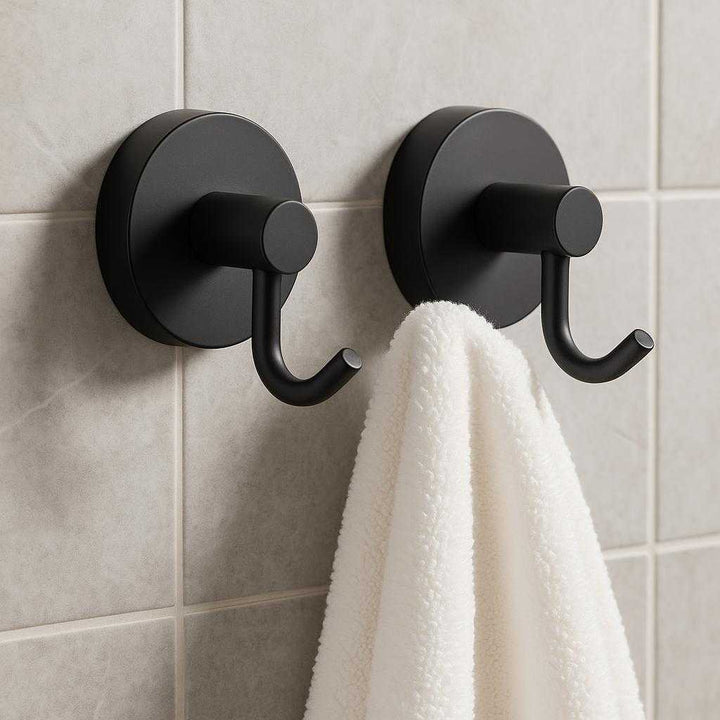 Vægkroge med sugekop - Sorte, selvklæbende kroge i moderne design til nem ophængning (2 stk): A realistic modern bathroom setting where the product is properly grounded on a smooth, tiled wall, being actively used to hold a fluffy white towel, with natural lighting casting realistic shadows and showing clear contact points where the hooks adhere to the wall.