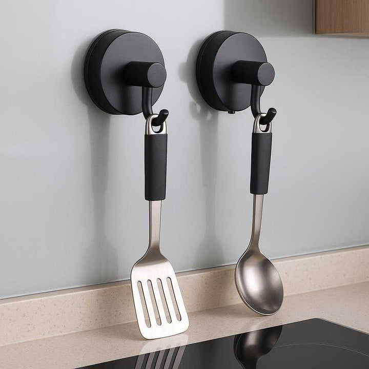 Vægkroge med sugekop - Sorte, selvklæbende kroge i moderne design til nem ophængning (2 stk): A different realistic kitchen setting where the product is securely positioned on a sleek glass backsplash, being used to hang a set of cooking utensils, in a believable way, with proper physics, shadows, and realistic integration into the environment.